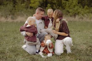 Family of five posing outdoors on green grass, parents holding young children in autumn-toned outfits near trees.