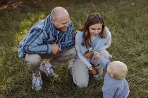 Family of three enjoying outdoor time together, with grandparents crouching down to interact with a toddler on green grass.