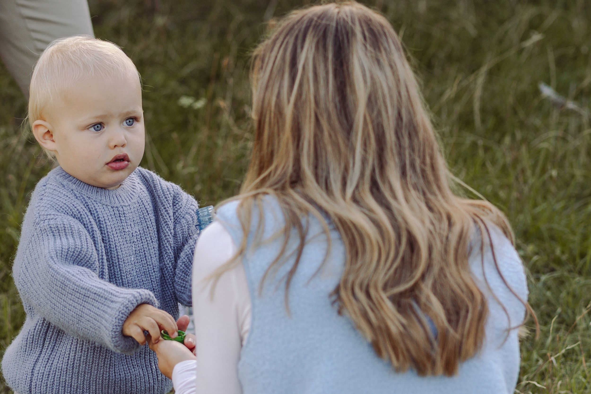 A toddler in a gray knit sweater interacts with a woman with long blonde hair sitting in a grassy field.