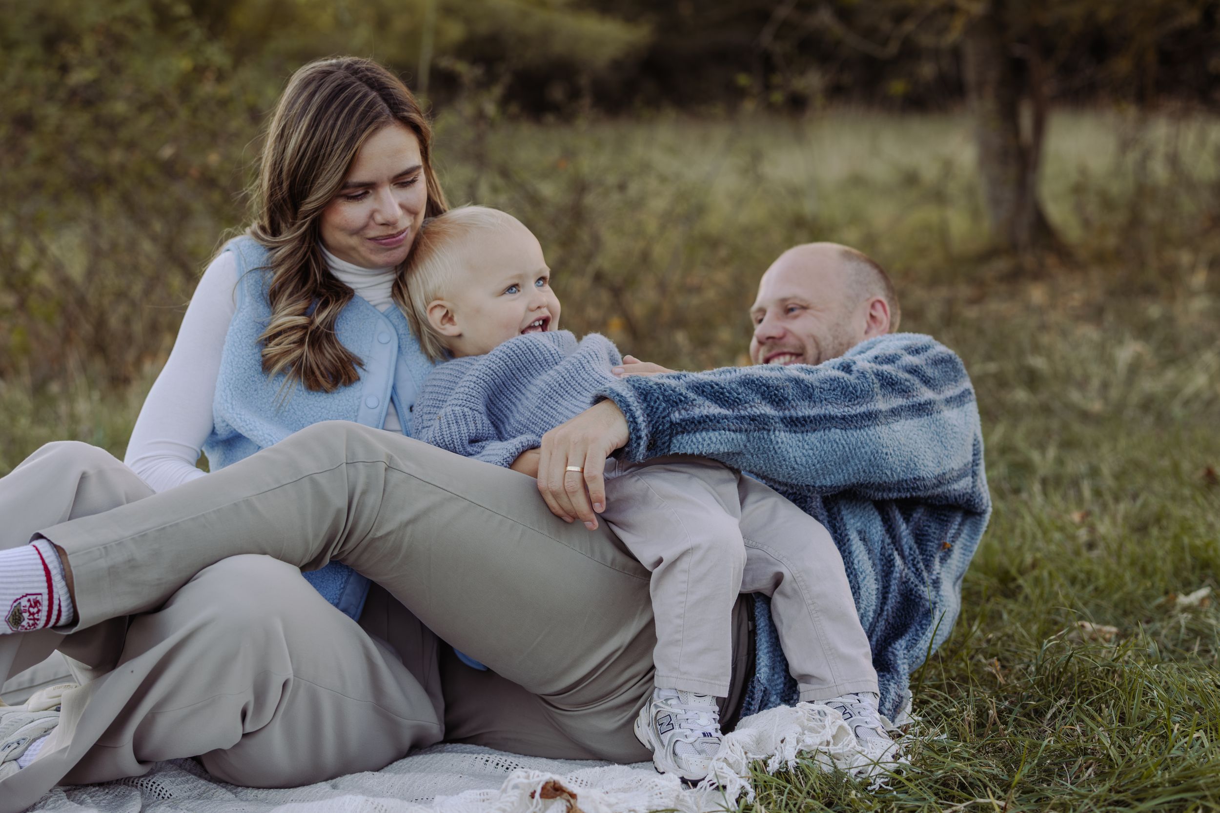 Happy family with baby relaxing on blanket in autumn park, parents smiling at laughing toddler outdoors.
