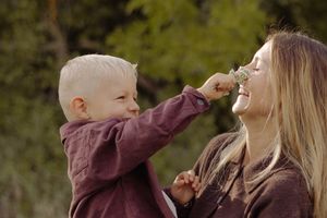 A young blond toddler holds a small flower to a smiling woman's nose outdoors in a green natural setting.