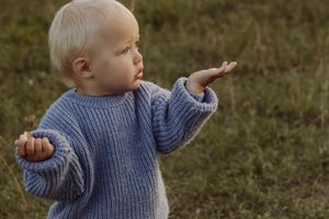 A blonde toddler in a blue knit sweater stands on grass with arms outstretched, looking curiously to the side.