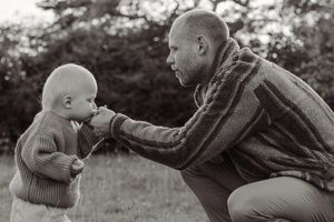 Black and white photo of a bald father crouching to interact with his toddler outdoors in a natural setting.