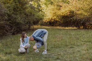 A couple plays together in a lush green field surrounded by autumn trees during an outdoor photo session.