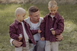 A father crouches with his two young blonde sons outdoors, all wearing matching burgundy shirts in a sunny field.