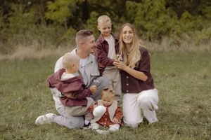 Happy family of four sitting on grass in a park, parents smiling with two young children in coordinating burgundy outfits.