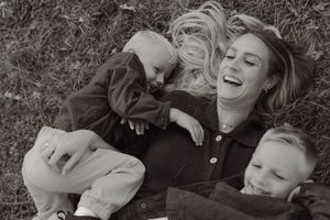 A laughing woman lies on grass with two young children in a joyful black and white outdoor family photo.