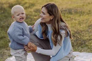 A smiling woman in a blue sweater sits outdoors holding a laughing toddler in a matching blue knit sweater.