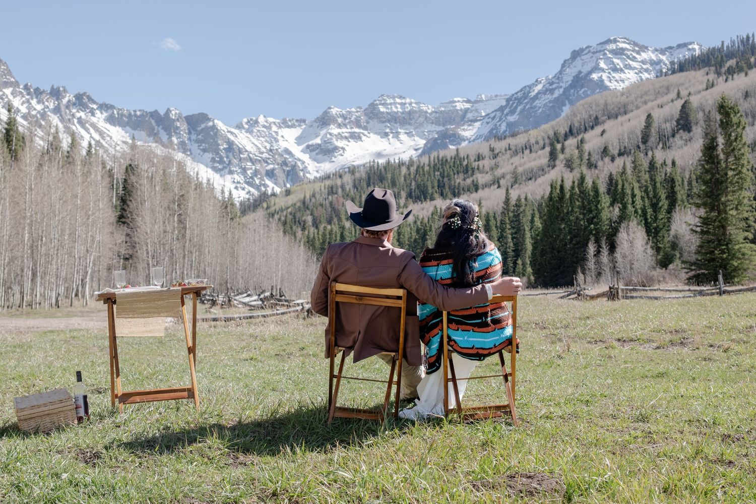 An eloping couple sitting in wooden chairs admiring snow-capped mountain views in a scenic alpine meadow with evergreen trees.