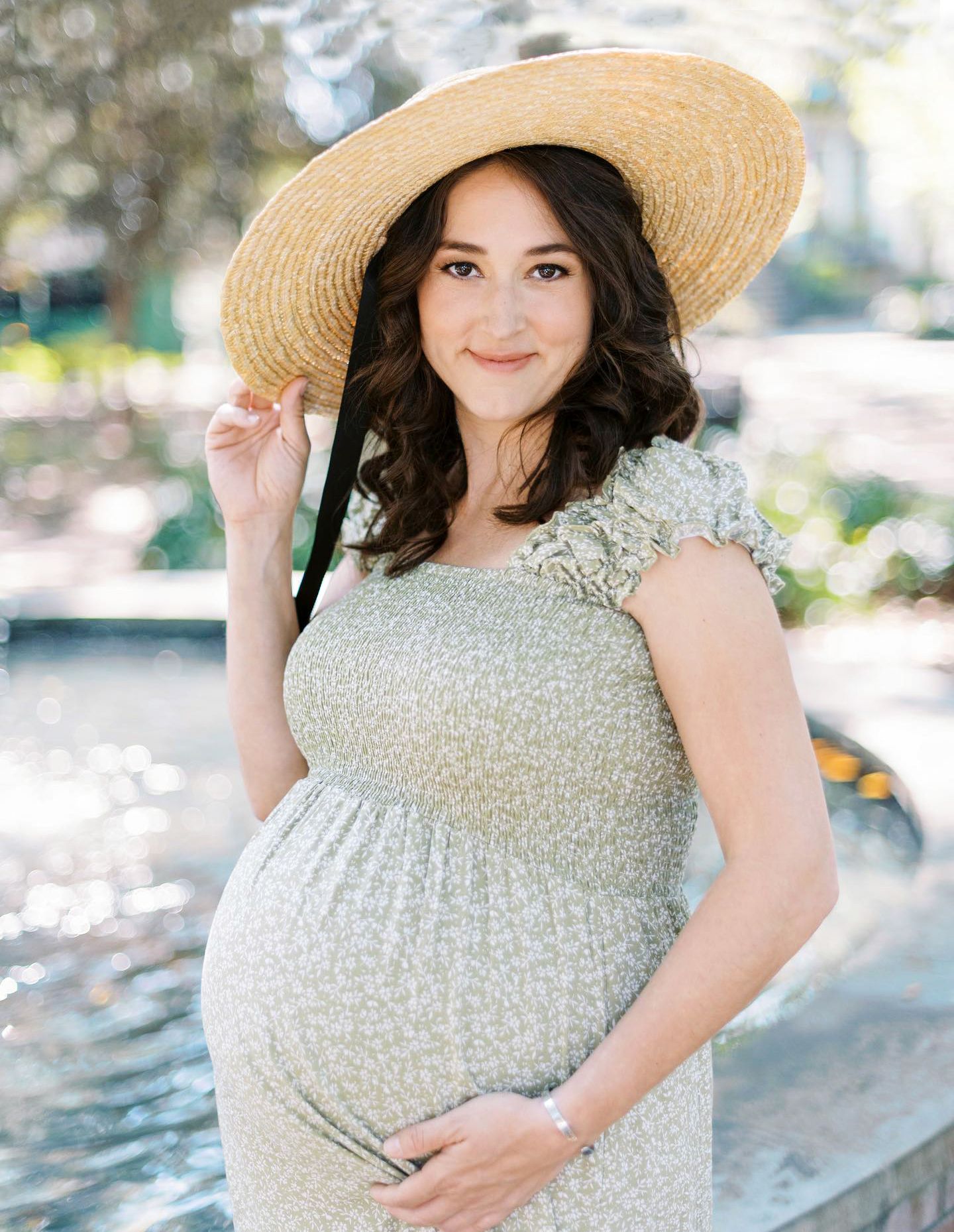 Pregnant woman in floral dress and wide-brim straw hat poses outdoors near fountain for maternity photo session.