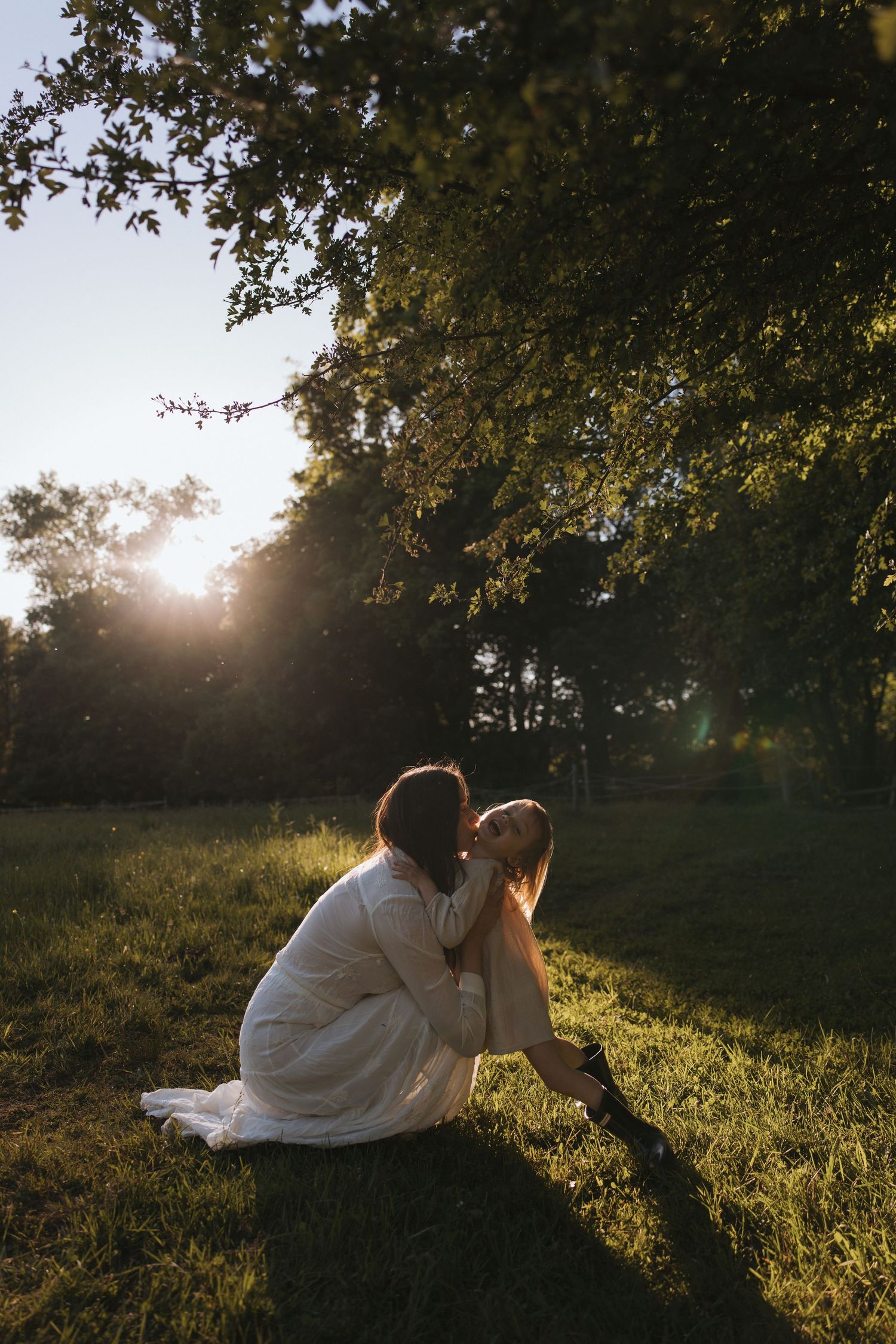 A mother tenderly kisses her child while sitting on lush green grass during golden hour sunset in a serene park.