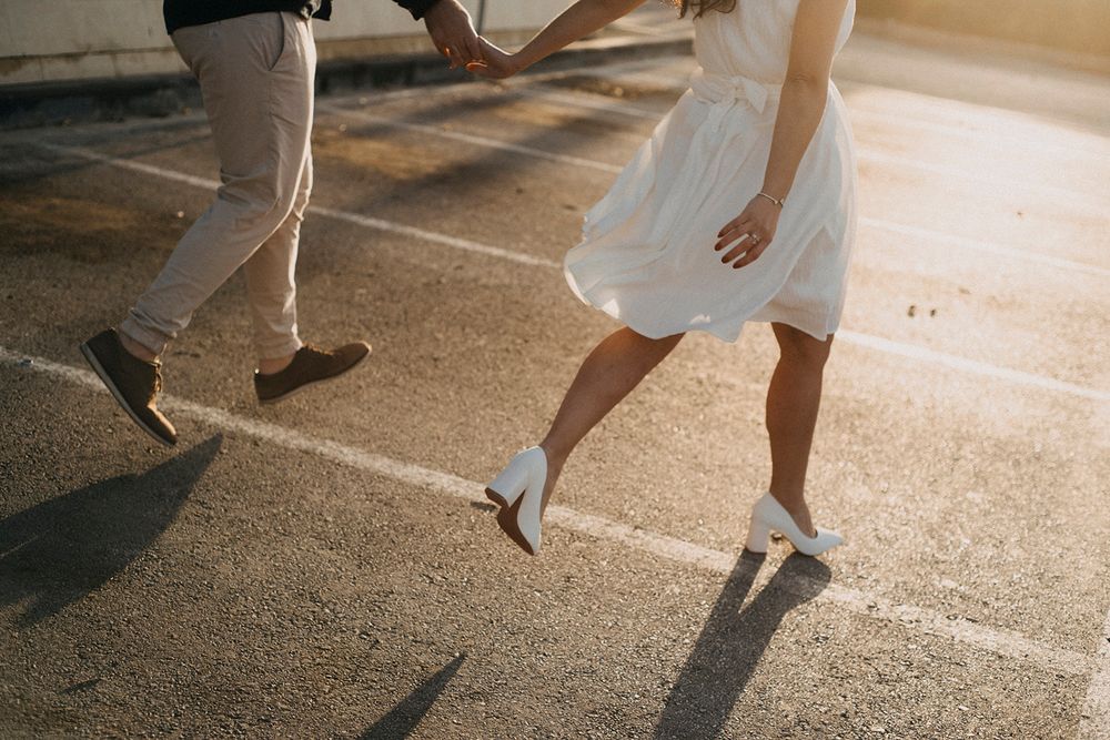 Couple holding hands running in a sunlit parking lot, woman in white dress and heels, man in casual pants and loafers.