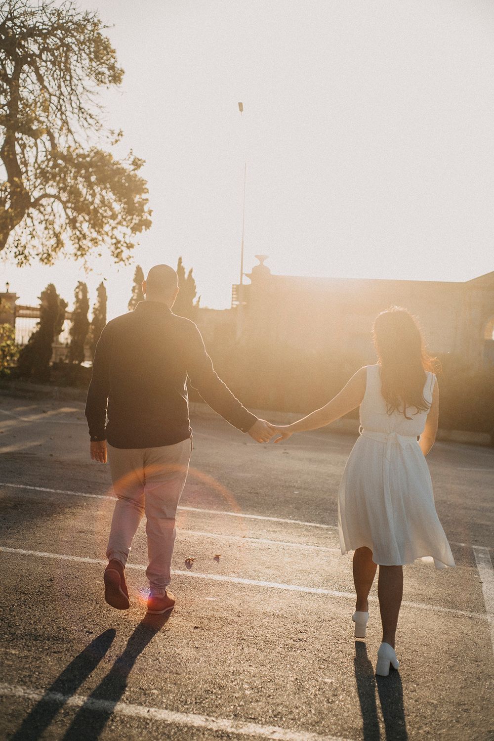 Couple holding hands walking into golden sunset light in a parking lot, backlit romantic photo.