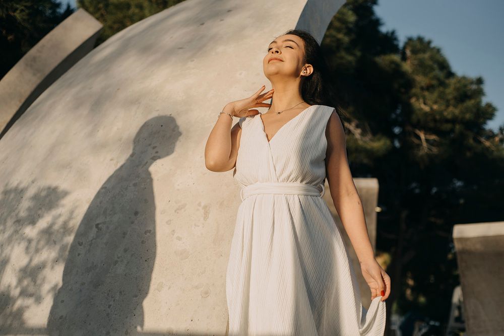 Woman in white dress poses gracefully outdoors near concrete sculpture with trees in background.