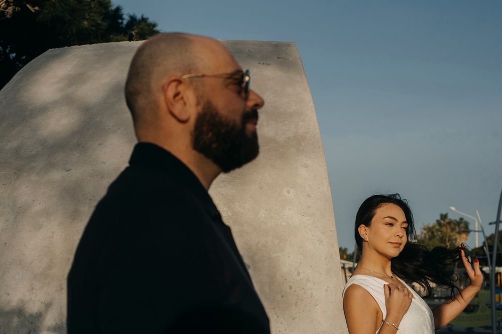 A bearded groom in black stands near a stone sculpture as a bride in white looks away during golden hour.