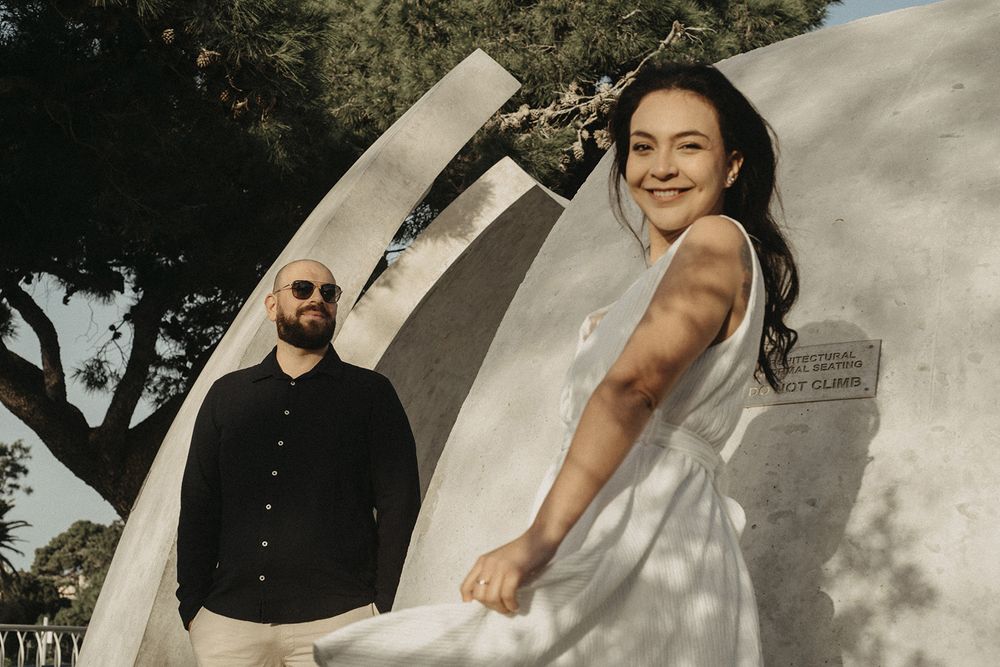 Couple in white and black outfits posing together near large concrete sculpture outdoors.