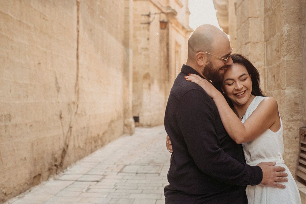 Couple embracing in a narrow stone alley, woman smiling in white dress, man in black outfit, warm golden tones.