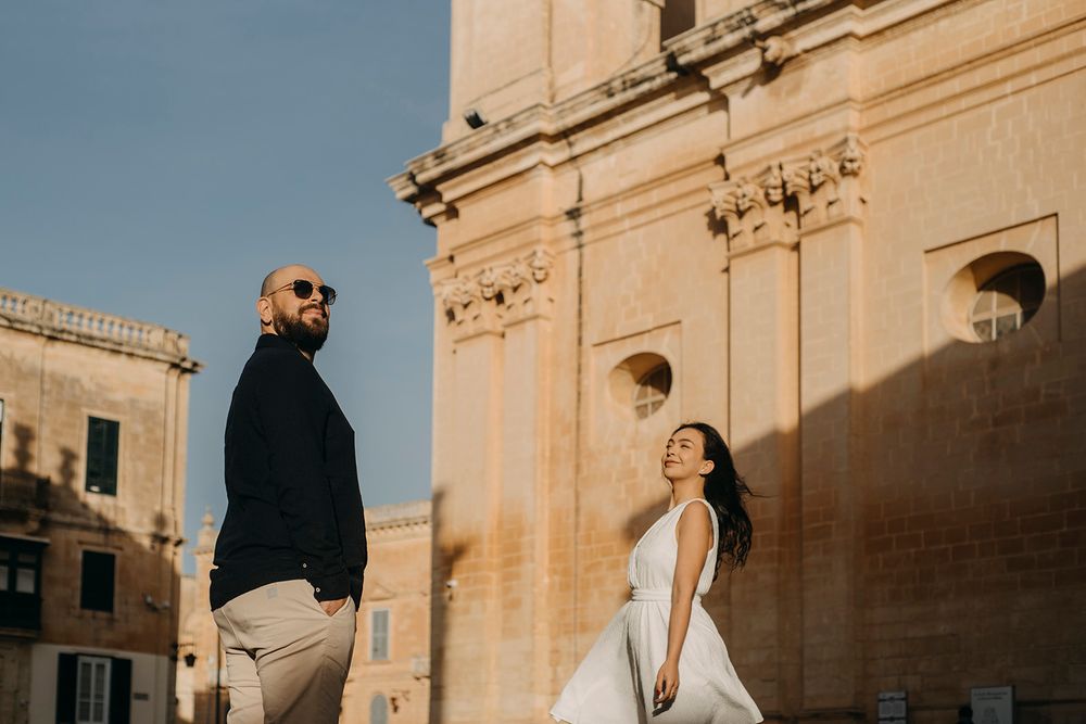 Couple in wedding attire pose near a historic stone building in Malta, bride in white gown, groom in black jacket.