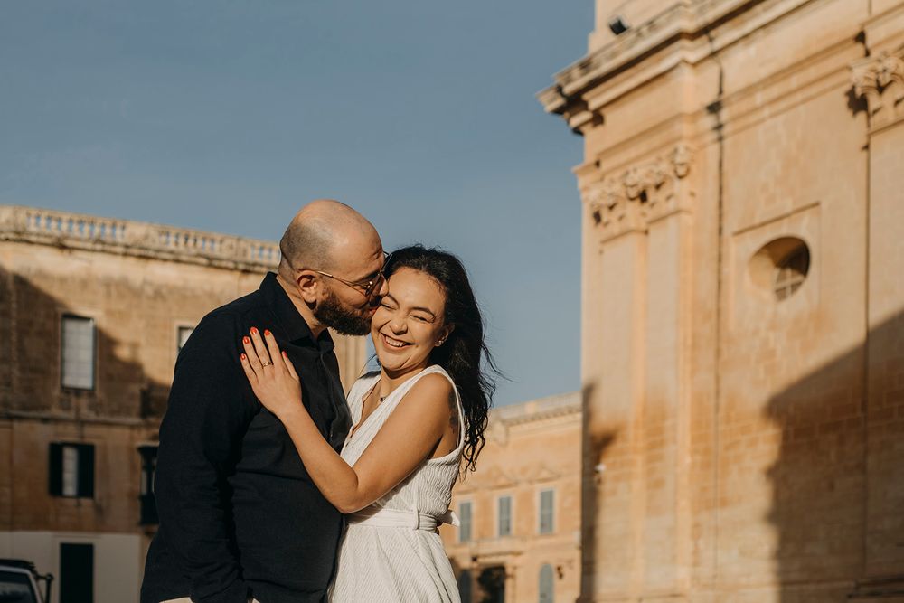 Happy couple sharing a tender moment near ornate stone architecture, woman in white dress smiling, man in dark suit.