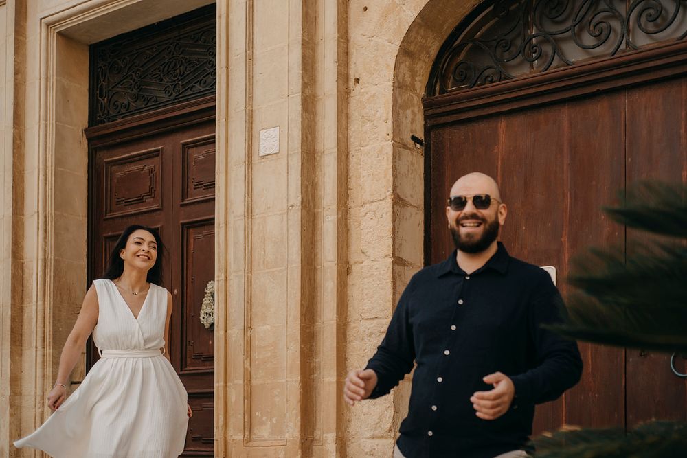 Smiling couple in white dress and black shirt walking past ornate wooden doors in a Mediterranean stone building.