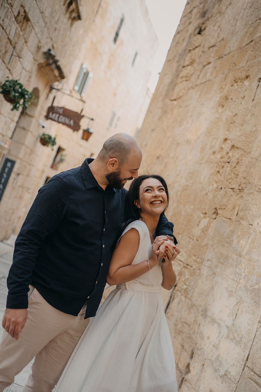 Couple walking hand in hand through a charming stone alley, smiling joyfully during their romantic engagement shoot.