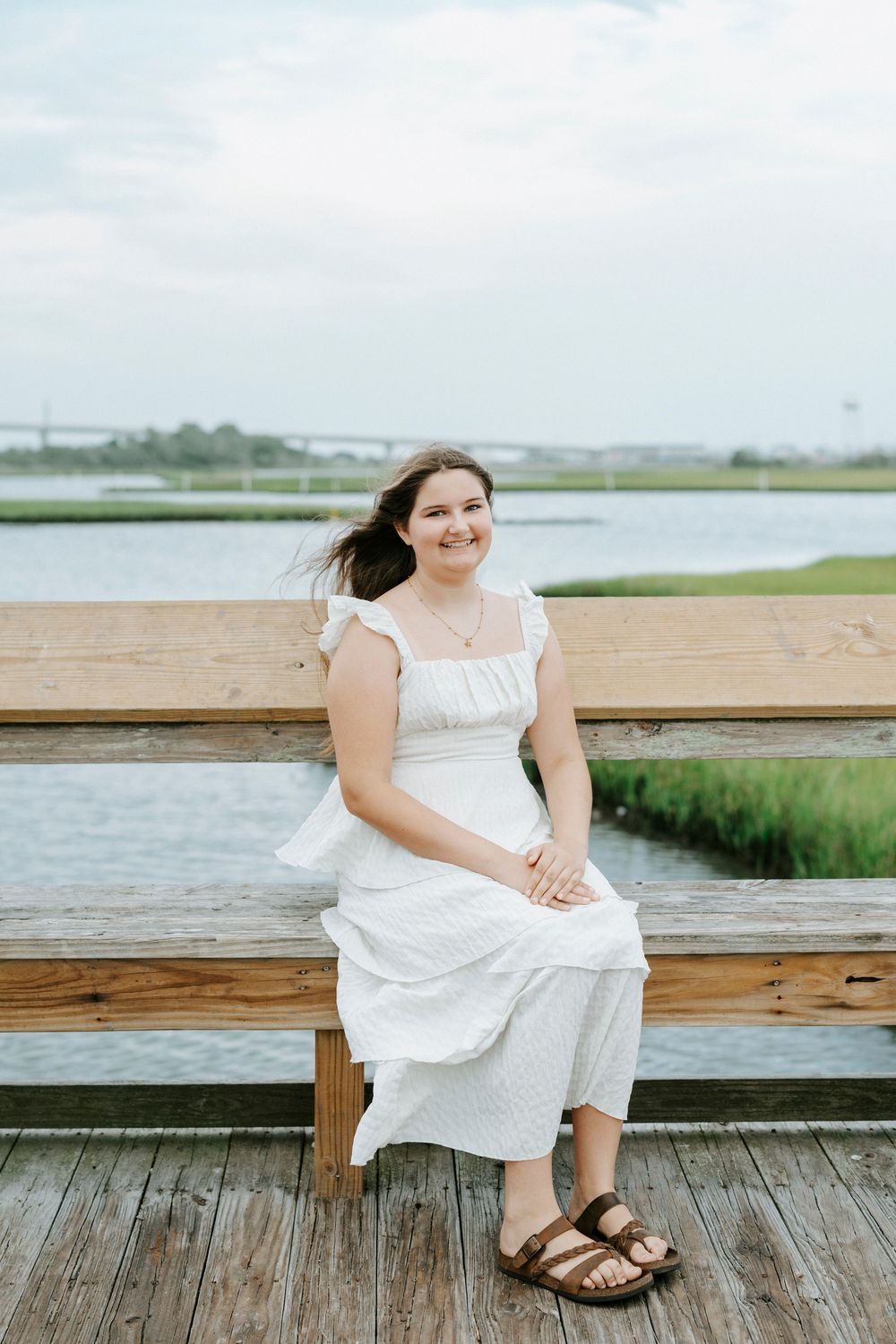 Young woman in white dress sitting on wooden dock bench by waterway, smiling outdoors.
