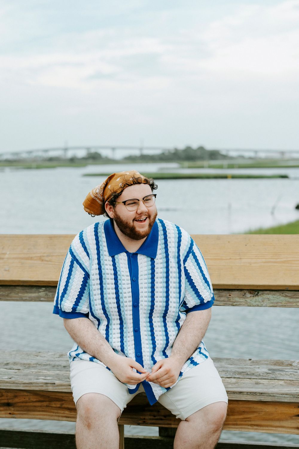 Man in blue striped crochet shirt and bucket hat sitting on wooden bench near waterfront, smiling outdoors.