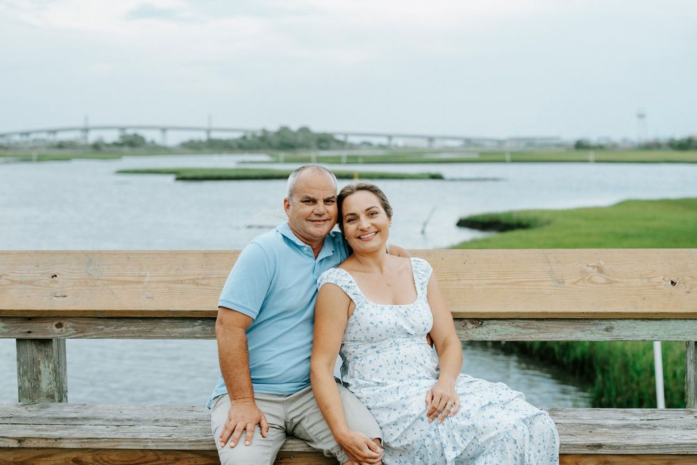 Couple smiling together on a wooden bench by a scenic waterway with green marshes in the background.