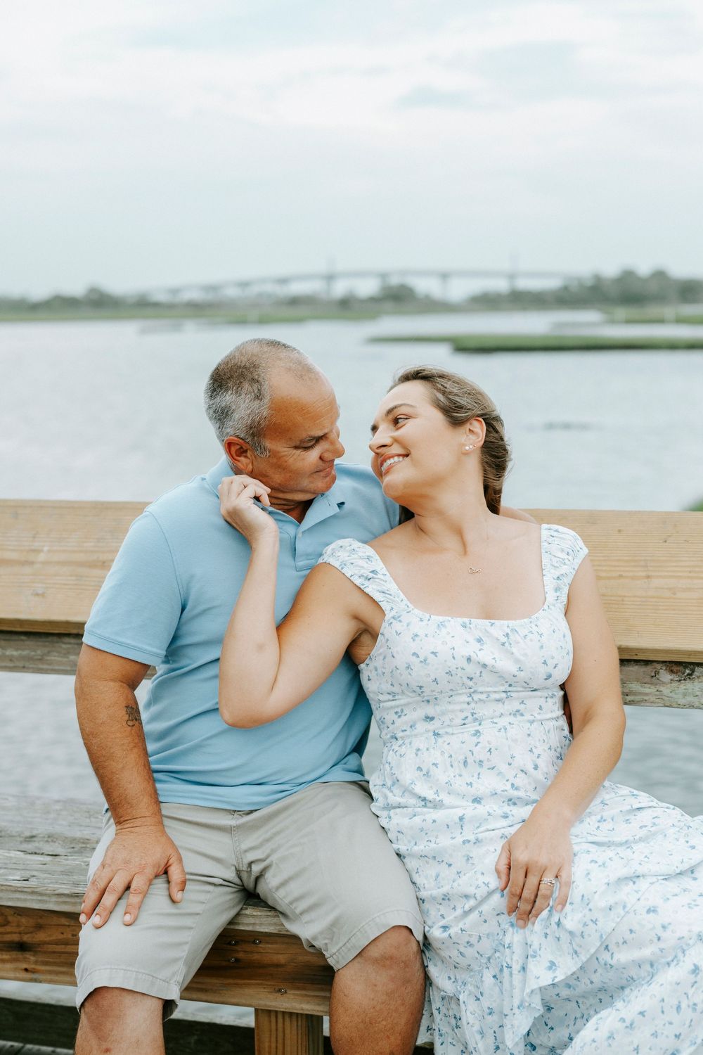 Couple sitting on wooden dock bench by water, man kissing woman's shoulder as she smiles softly.