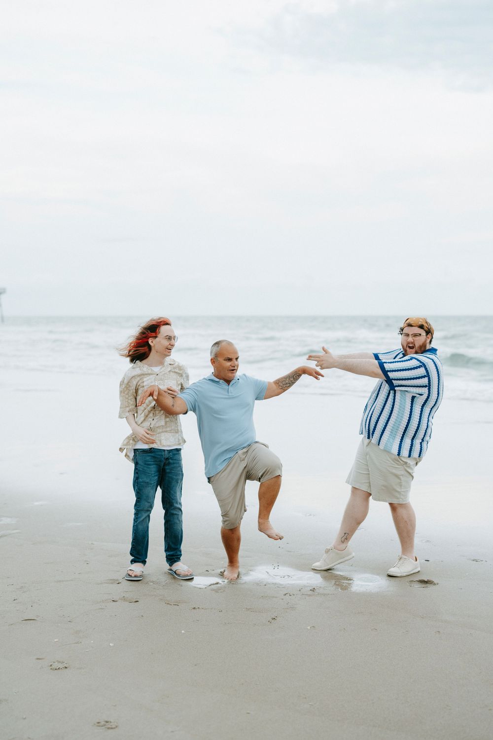 Three friends playfully posing on a sandy beach with ocean waves in the background on a cloudy day.