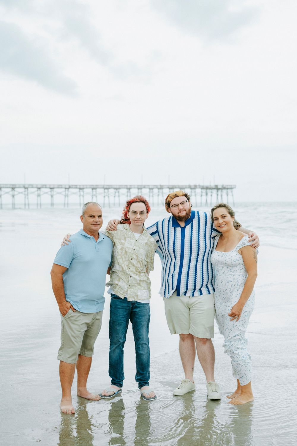 Family of four posing together on a beach near a pier, smiling on a cloudy day.