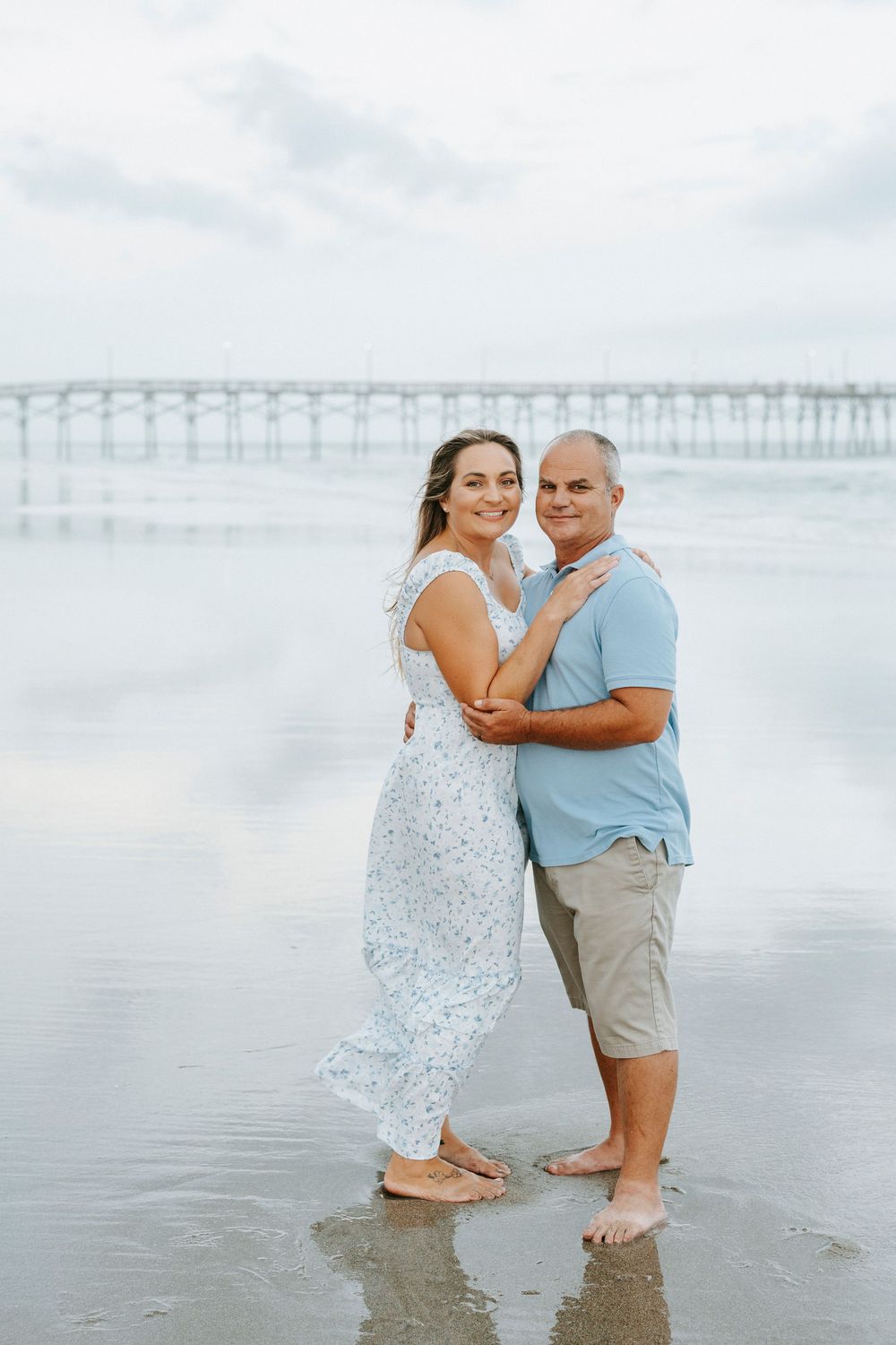 Couple embracing on a reflective beach with a pier in the background on an overcast day.