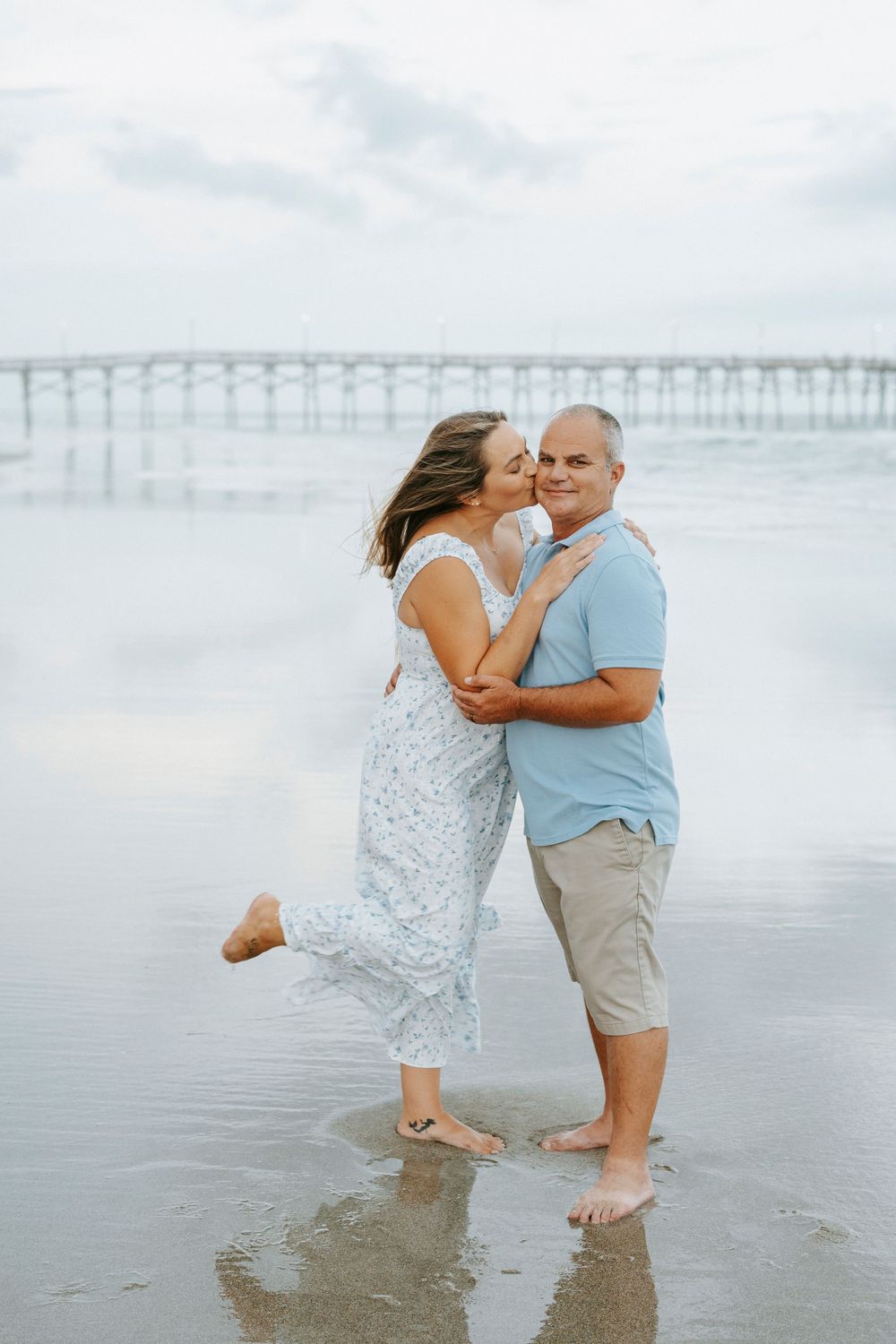 Couple sharing a romantic kiss on the beach near a pier, woman in floral dress lifting one leg.