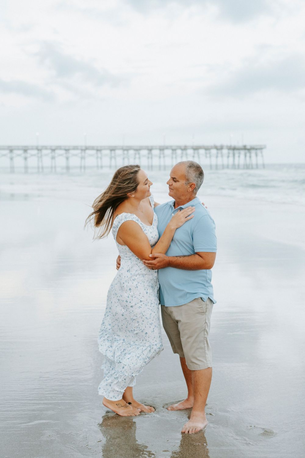 Couple dancing barefoot on a reflective beach shore with a pier in the background on a cloudy day.