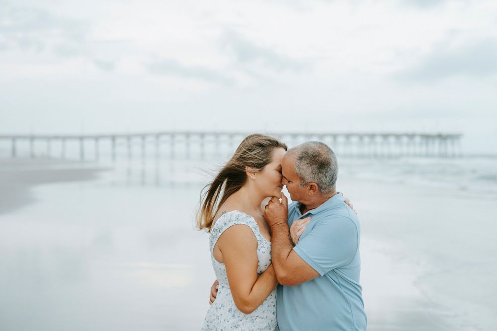 Romantic couple embracing on a beach near a pier, sharing a tender moment with ocean waves in the background.
