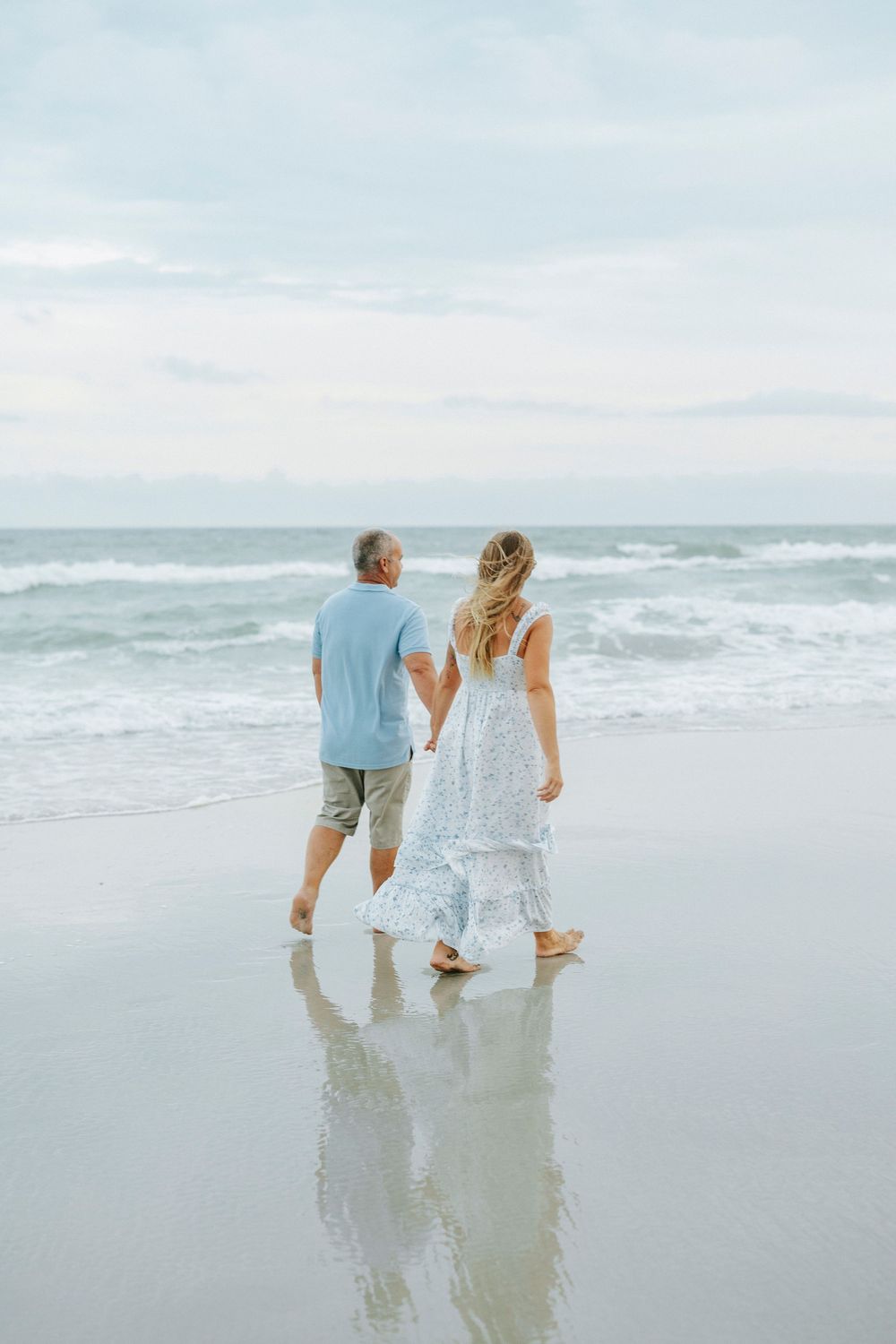 Couple holding hands walking barefoot toward ocean waves on a calm, cloudy beach day.