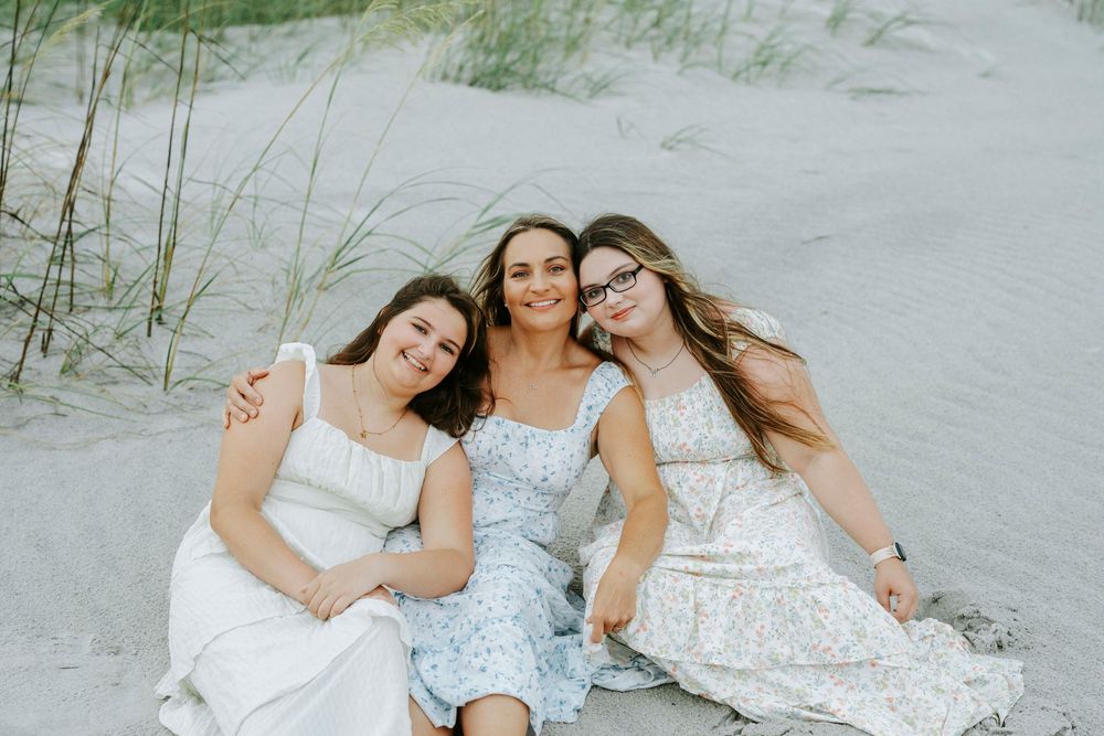 Three young women in white and floral dresses smile together on a sandy beach near grass-covered dunes.