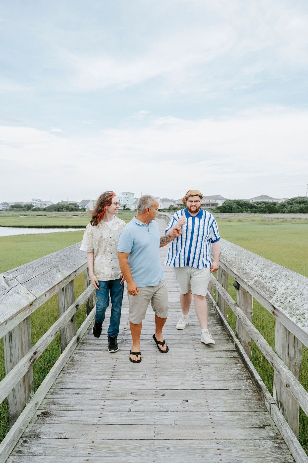 Three people walking together on wooden boardwalk over marsh grasslands under cloudy sky.
