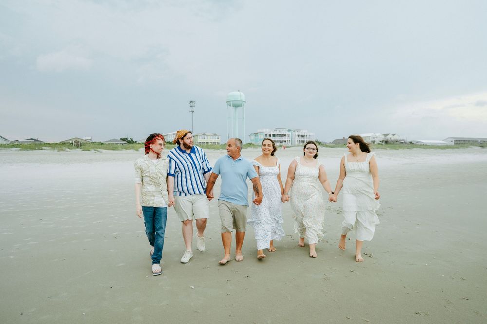 Six people walking hand-in-hand on a beach with a water tower in the background on a cloudy day.