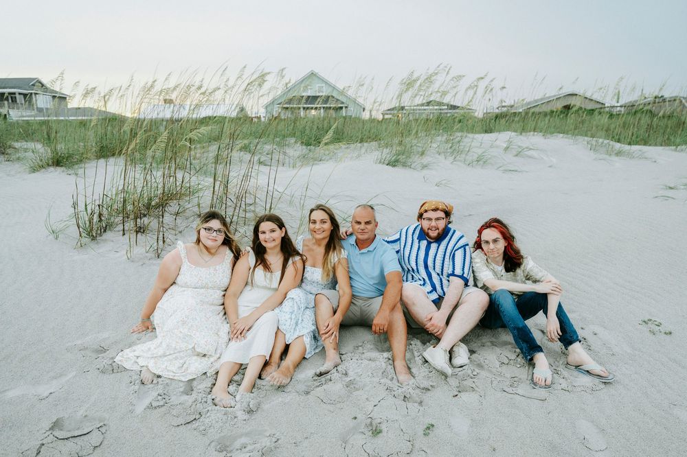 Family of six smiling together on a sandy beach with sea oats and colorful beach houses in the background.