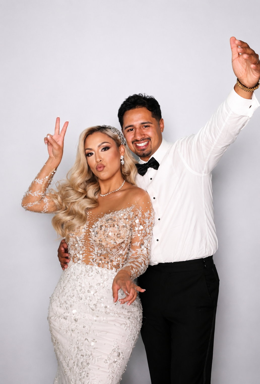 A bride in a beaded gown and groom in a tuxedo pose playfully, flashing peace signs at a wedding photo booth.