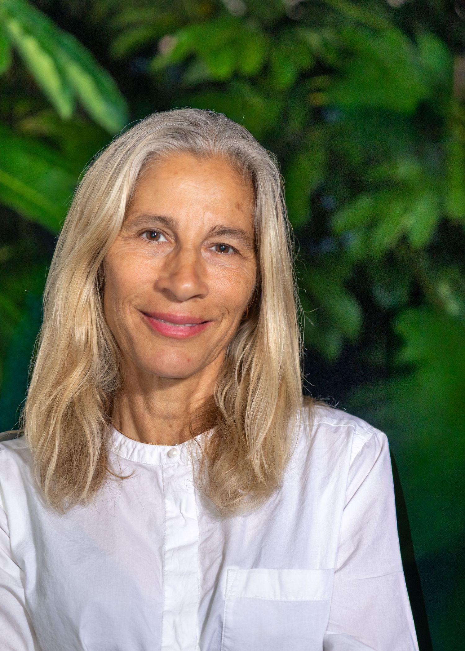 Older woman with gray hair holding a DSLR camera, wearing a striped shirt, posing in front of a pink curtain.
