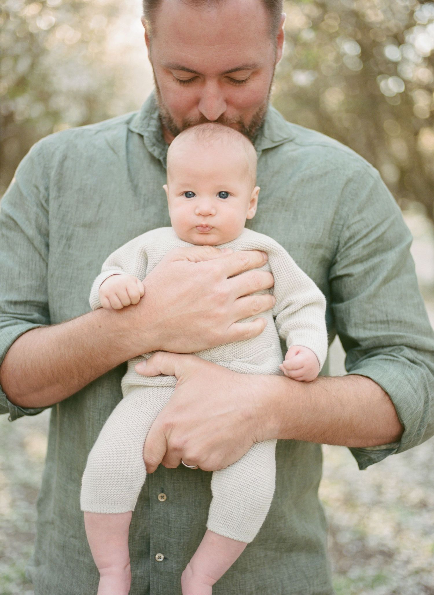 A bearded father in a green shirt kisses his infant baby's head while gently holding the child outdoors.
