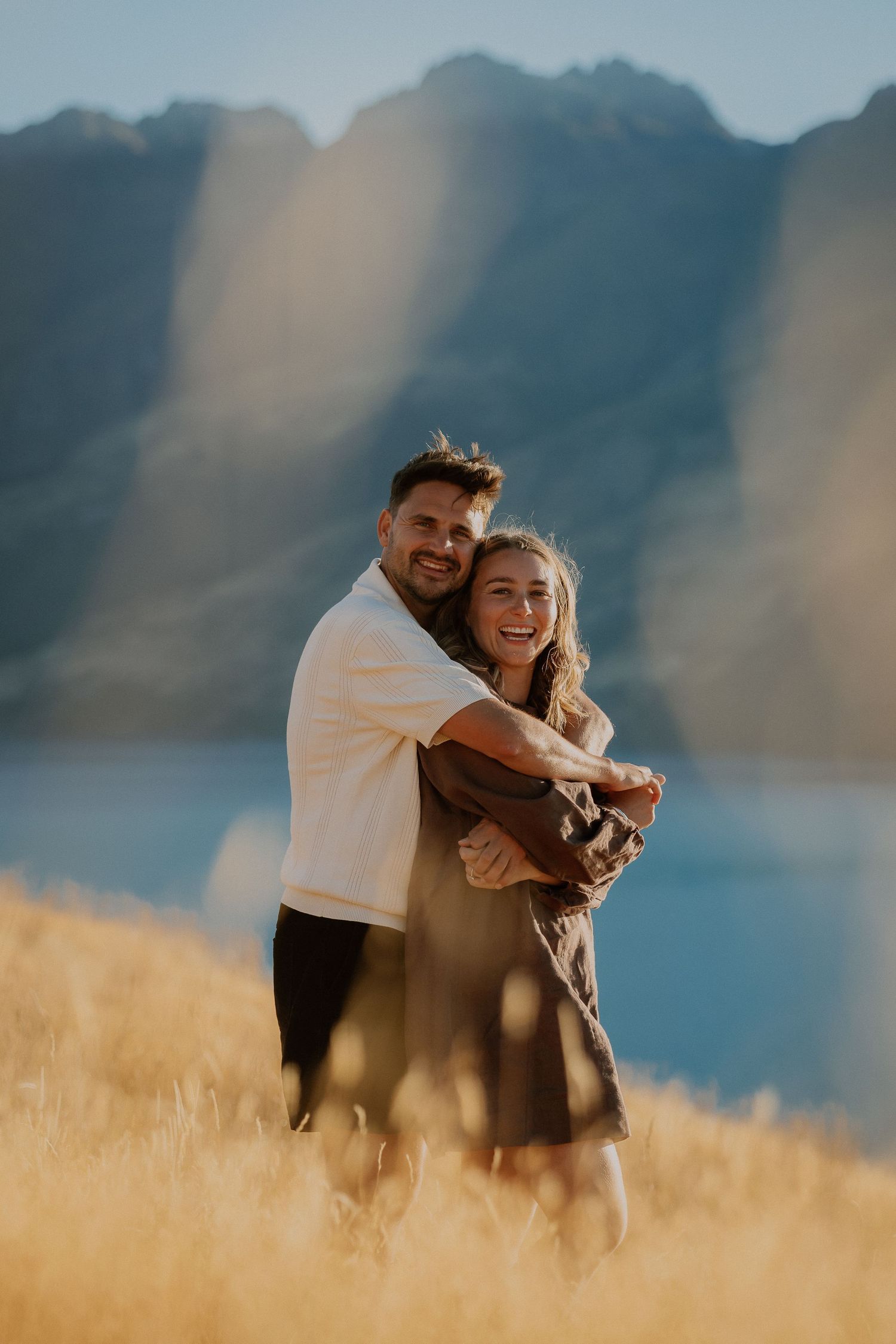 Couple embracing romantically on golden hillside with blue lake and mountains in background on sunny day.