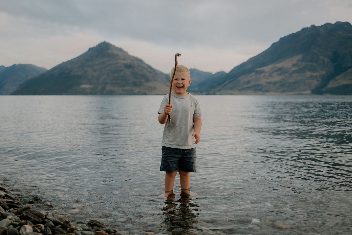 Young boy standing in a lake holding a stick with mountains in the background on a cloudy day.
