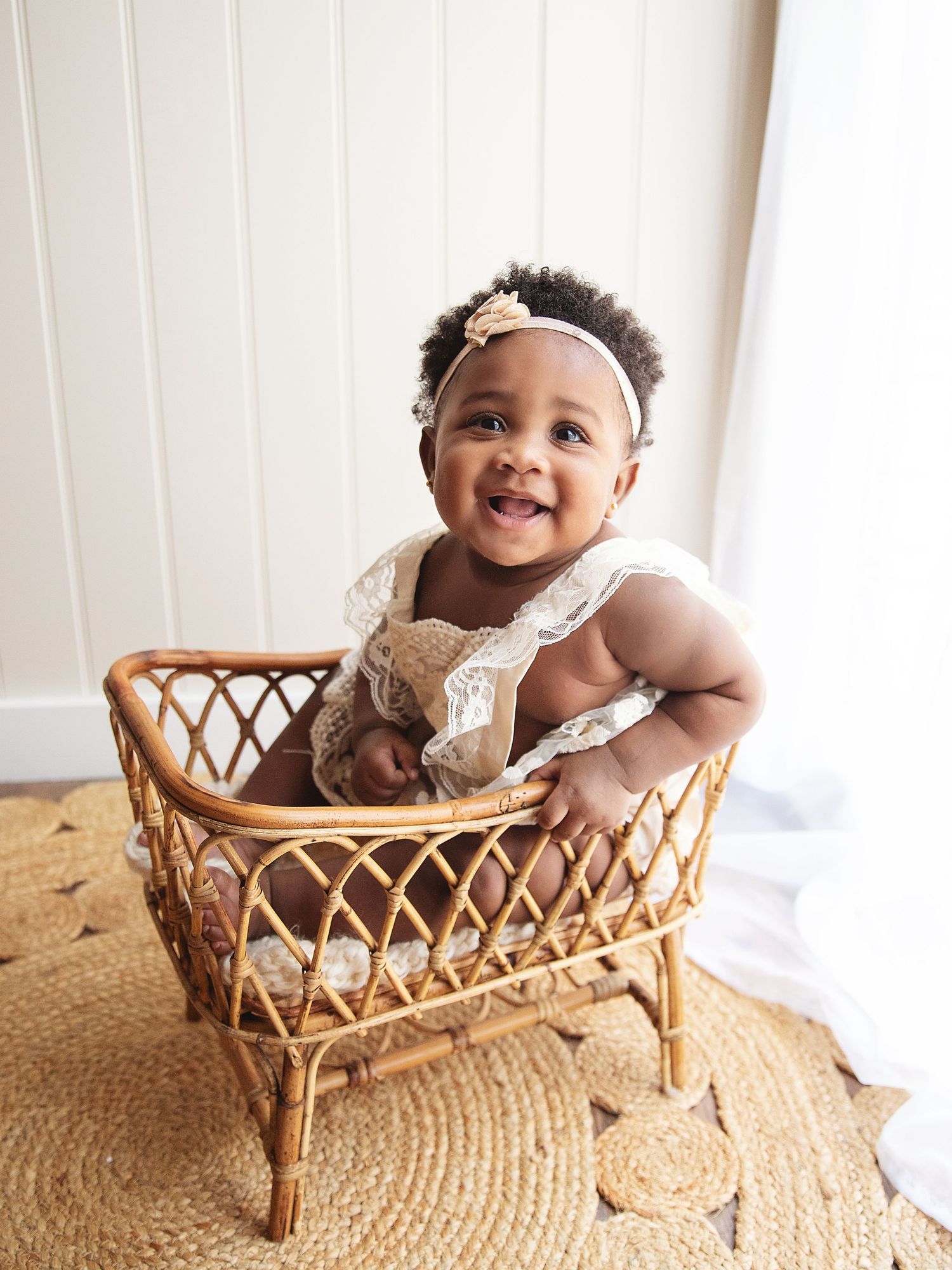 Smiling baby girl in white lace outfit and floral headband sitting in a rattan bassinet. During a 6 month baby photoshoot in Sidcup