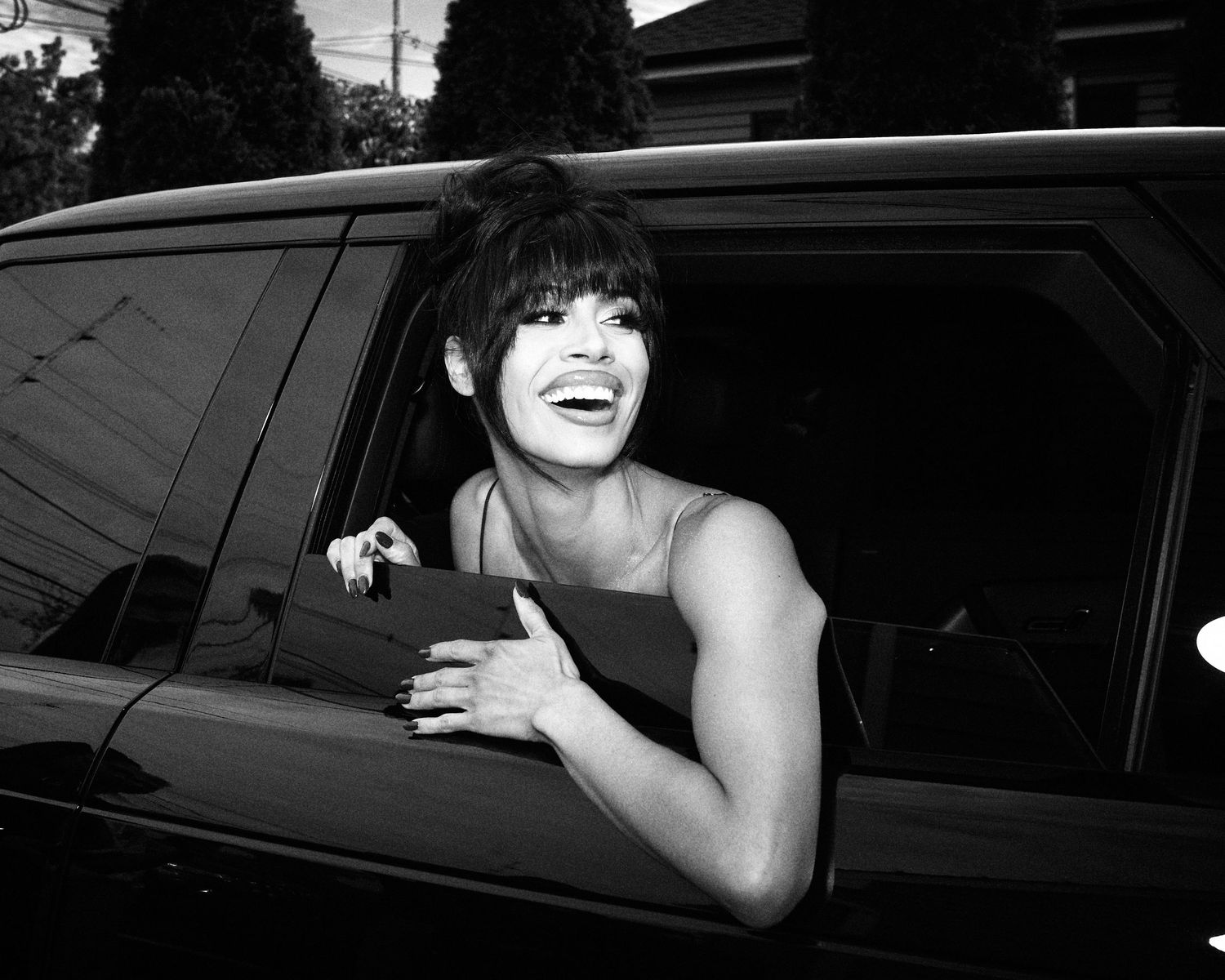 Black and white photo of a laughing woman leaning out of a car window, wearing a sleeveless top.