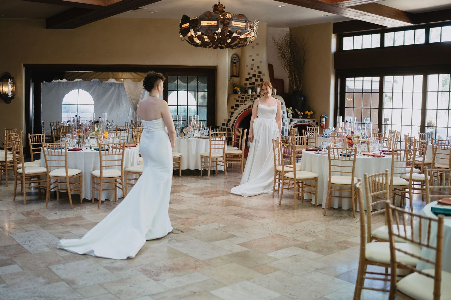 Two brides in elegant white gowns pose in a beautifully decorated wedding reception hall with gold chairs.