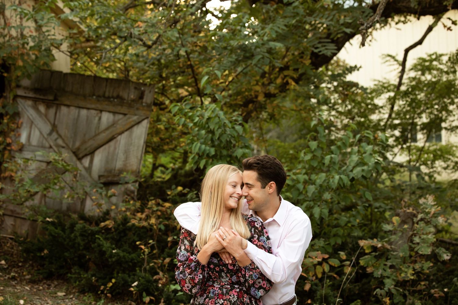 A couple embraces and smiles together outdoors surrounded by lush green trees and foliage near a rustic wooden gate.