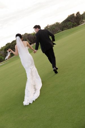 Newlyweds holding hands running across a lush green golf course in wedding attire.